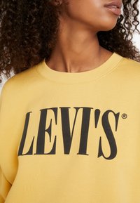 Femme aux cheveux bouclés portant un sweat-shirt jaune avec un grand texte noir "LEVI'S" sur la poitrine, regardant sur le côté.
