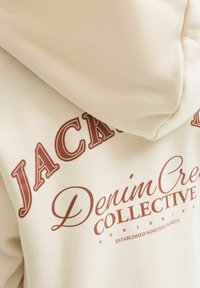 Felpa beige con testo marrone che recita "JACK & DENIM CREW COLLECTIVE WORLDWIDE FONDATA NELL'ANNO NOVANTANOVE" sul retro.