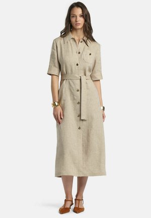 Kvinde iført en beige midi-kjole med korte ærmer, knapper foran, bælte i taljen og brune T-strap sko med spids snude, stående mod en hvid baggrund.