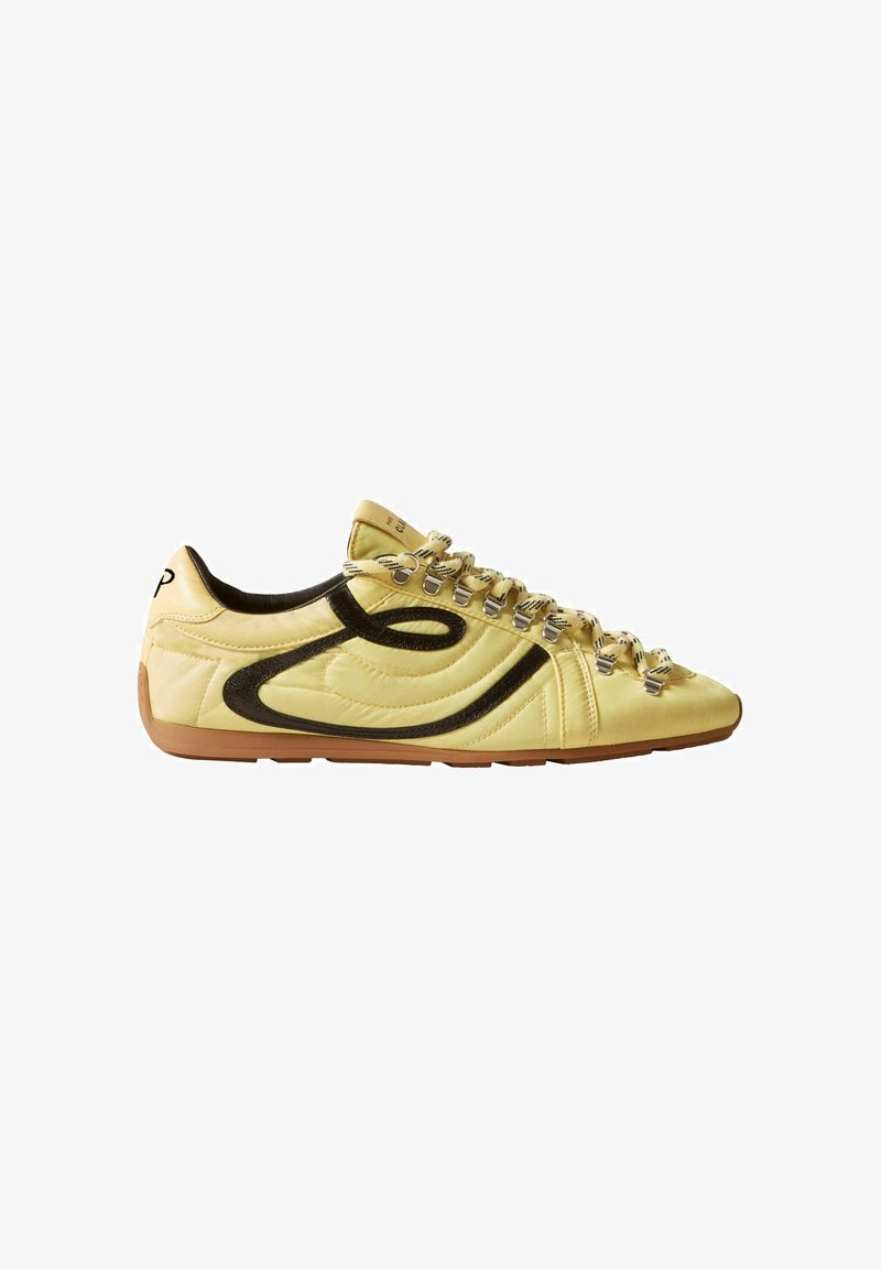 Zapatilla casual de color amarillo claro con suela marrón, diseño lateral ondulado negro y cordones beige pasados por ojales metálicos.