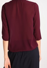 Blouse en voile bordeaux avec des manches trois-quarts, un col froncé et un ourlet élastiqué. Texture lisse, tissu léger et coupe classique.