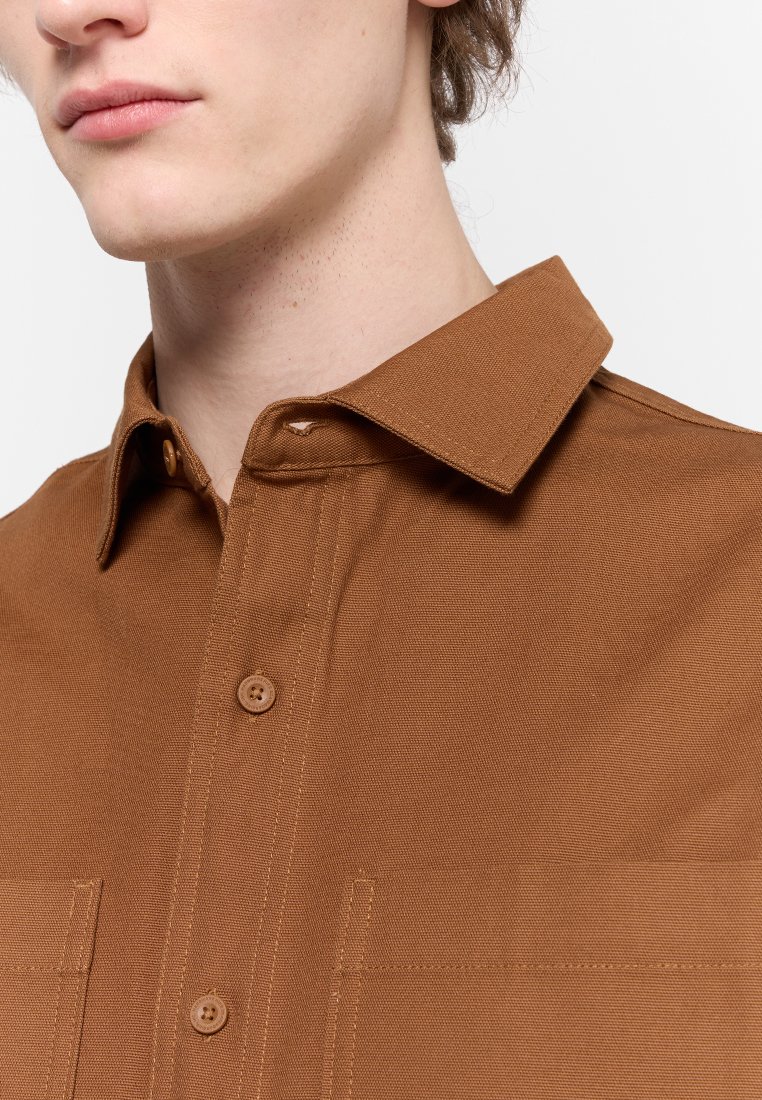 Jeune personne portant une chemise brune à boutons avec un col pointu, présentant la partie supérieure de la poitrine et le cou sur un fond blanc.