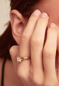 Bague en or avec une pierre précieuse transparente centrale, présentant un anneau lisse et des accents texturés. Gros plan d'une main couvrant partiellement le visage.