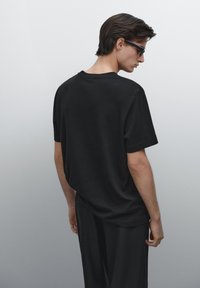 Schwarzes Baumwoll-T-Shirt mit lockerer Passform und kurzen Ärmeln, das einen klassischen Rundhalsausschnitt und ein minimalistisches Design aufweist, von hinten betrachtet.