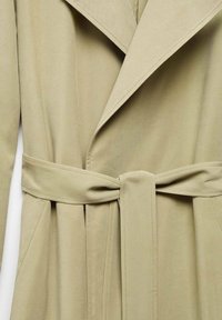 Trench coat beige chiaro con colletto a punta, maniche lunghe e vita con cintura. Il tessuto ha una texture liscia e presenta una silhouette su misura.