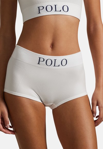 Kobieta ubrana w białe prążkowane szorty z wysokim stanem i dopasowany krótki top, oba z elastycznymi taśmami z logo "POLO", stojąca na jednolitym tle.
