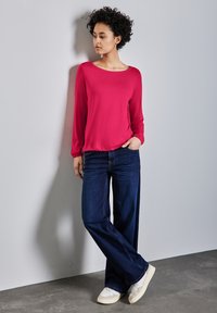 Langärmliges fuchsia-farbenes Oberteil, kombiniert mit weiten, dunklen Jeans, die eine lockere Passform und eine weiche Textur bieten. Weiße Sneaker runden das Outfit ab.