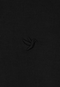 Sort stof med en ribbet struktur, der præsenterer et diskret broderet kolibrilogo, også i sort, placeret i nederste venstre hjørne.