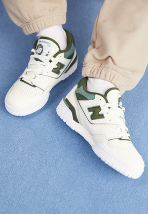 Bílé tenisky New Balance 550 se zelenými akcenty, nošené s bílými ponožkami a béžovými kalhotami na modrém koberci.