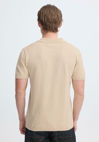 Man met kort bruin haar, een beige shirt met korte mouwen en donkere jeans, staand met de rug naar de camera tegen een egale lichte achtergrond.