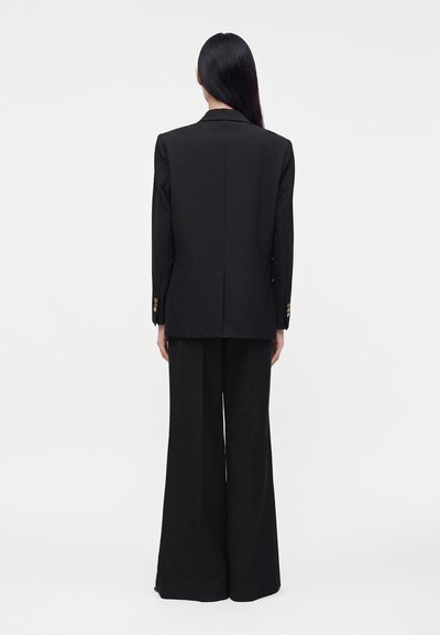 Femme aux longs cheveux foncés portant un blazer noir et un pantalon large noir, debout de dos devant un fond clair uni.