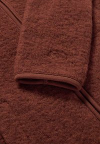 Braunes Fleece-Material mit strukturierter Oberfläche, featuring einen gerippten Saum und einem dezenten Logo-Detail. Reißverschluss entlang einer Kante.