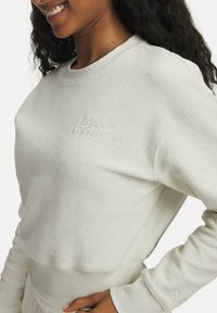Sweatshirt beige clair avec texture côtelée, col rond et logo brodé sur la poitrine. Présente des manches longues et un design court.