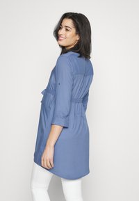 Tunic a maniche lunghe, di colore azzurro chiaro, con cintura in vita, caratterizzata da un orlo curvo e un tessuto morbido, che mette in mostra una silhouette rilassata.