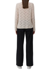 Femme de dos face à la caméra, portant un pull en tricot beige avec un motif en dentelle, un jean noir à jambe large et des baskets blanches.