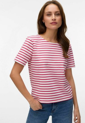 Kvinde med brunt hår iført en hvid T-shirt med korte ærmer med vandrette røde striber og blå jeans, poserende med den ene hånd i lommen.