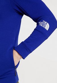 Modrá mikina se zipovou kapsou, vyrobená z měkké látky. Na rukávu je bílý logo a manžety jsou žebrované pro pevné uchycení.