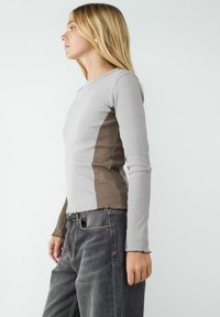 Lange mouwen top van lichtgrijze ribstof, met een donkerbruine paneel aan de zijkant. De zoom is licht gegolfd. Gecombineerd met donkere jeans.