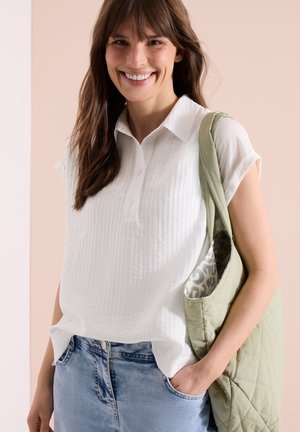 Glimlachende vrouw met lang bruin haar draagt een wit kort mouwtjes blouse met knopen, lichtblauwe jeans en draagt een lichtgroene gewatteerde shopper tas.