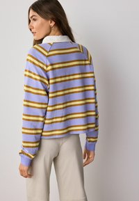 Donna con i capelli lunghi e castani, che indossa un maglione a strisce lavanda e senape con colletto bianco e pantaloni beige chiaro, girata di lato.