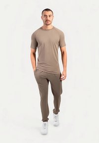 Beige T-shirt met korte mouwen en een ronde hals, gecombineerd met bijpassende taps toelopende broek. Het model draagt witte sneakers en staat tegen een neutrale achtergrond.