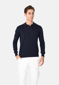 Polo a maniche lunghe blu marina realizzato in un tessuto morbido, con colletto classico e pattina con tre bottoni, indossato con pantaloni bianchi.