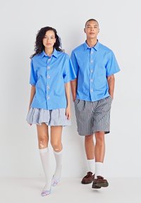 Lyseblå skjorter med blomsterdetaljer, kombinert med et stripete skjørt og shorts. Modellen har på seg hvite knelange sokker og sandaler.