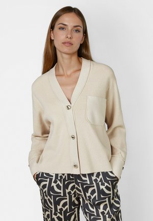 Woman wearing a beige buttoned cardigan with chest pocket and black and beige patterned pants, standing with hands in pockets against white background.