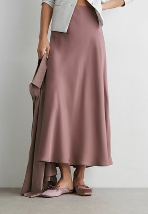 Kobieta ubrana w mauve satynową midi spódnicę, pasujące wsuwane baleriny, jasnoszarą krótka kurtkę i trzymająca mauve koszulę na guziki.