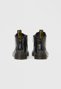 Μαύρα μποτάκια αστραγάλου με γυαλιστερό φινίρισμα, μεταλλικές πιτσιλιές, φερμουάρ στο πλάι και κίτρινες ταμπελίτσες με λογότυπο. Διαθέτουν παπούτσι από καουτσούκ με σχέδιο.