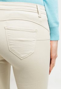 Jeans skinny beige con un bolsillo trasero en forma de diamante, hechos de un material de algodón suave, con detalles de costura sutiles.