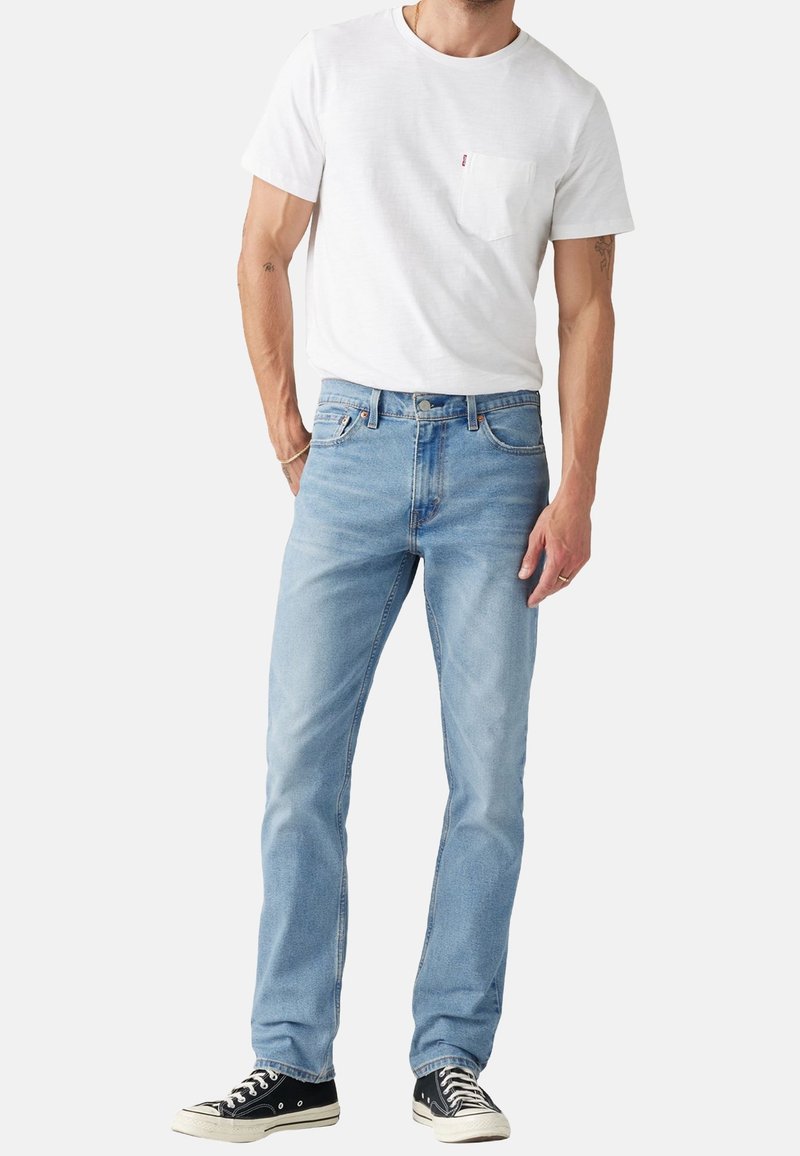 Homme portant un jean droit bleu clair, un t-shirt blanc à manches courtes avec poche et des baskets basses noires, debout devant un fond blanc.