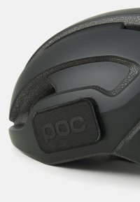 Casque de cyclisme noir avec un revêtement intérieur texturé, des ouvertures de ventilation et un patch logo POC sur le côté. Design aérodynamique et élégant.