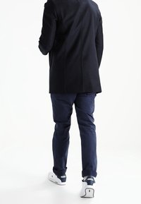 Persona con un abrigo negro, pantalones azul marino y zapatillas Lacoste blancas caminando hacia atrás sobre un fondo blanco.