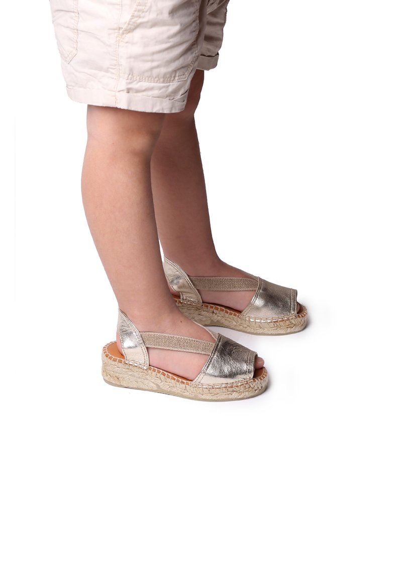 Metallische zilveren sandalen met open tenen, elastische banden en geweven jute zolen, vanaf de zijkant bekeken op de voeten van een kind.