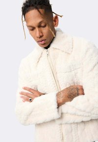 Homme avec des cheveux tressés portant une veste à zip blanche texturée, les bras croisés, montrant des tatouages et une bague à sa main droite.