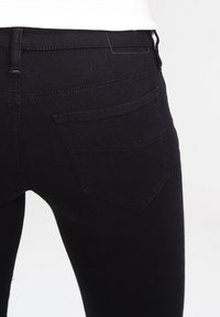 Jeans de mezclilla negra con un corte ajustado, que cuentan con un bolsillo trasero de diseño puntiagudo y detalles de costura sutiles. Textura suave y acabado mate.