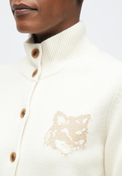 Közelkép egy olyan személyről, aki fehér kötött kardigánt visel, fából készült gombokkal és bézs színű rókafej mintával a mellkasán.