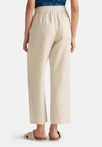 Beige Leinenhosen mit weitem Bein und elastischem Bund sowie einer einzelnen Gesäßtasche, von hinten gezeigt. Tragen mit flauschigen Slipper-Sandalen.