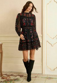 Fekete virágmintás ruha hosszú, áttetsző ujjal, derekán megkötve, fodros szegéllyel, piros és kék virágmintával, fekete csizmával párosítva.