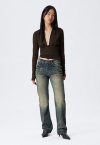 Hnědý dlouhý rukáv s hlubokým výstřihem do V, v kombinaci s vybledlými modrými džíny. Modelka má na sobě černé špičaté boty na podpatku. Jednoduchý, čistý design.