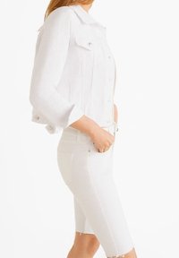 Chaqueta de mezclilla blanca con un diseño recortado, que cuenta con cierre de botones, dos bolsillos frontales y mangas enrolladas, combinada con pantalones cortos blancos a juego.