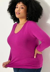 Top aderente a maniche lunghe in un vivace tessuto magenta con scollatura a V e superficie liscia. Abbinato a jeans blu con un leggero risvolto.
