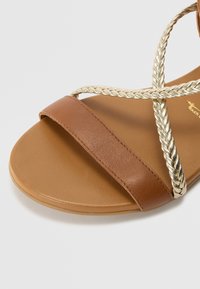 Sandalo piatto marrone con un'ampia fascia in pelle e un'acca di oro intrecciata. La suola presenta una finitura texturizzata per una buona aderenza e comfort.