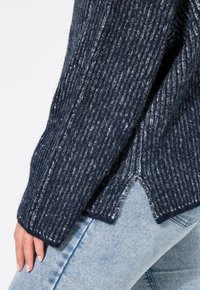 Marineblauer Strickpullover mit vertikaler Rippstruktur, seitlichem Schlitzsaum und kontrastierenden helleren Fasern. Kombiniert mit hellblauen Jeans.