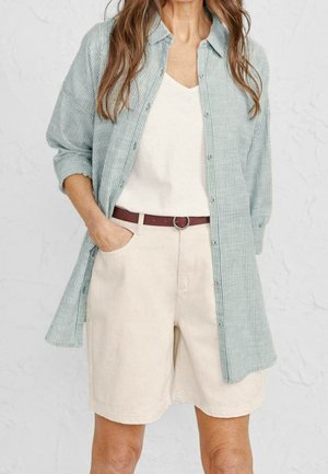 Femme portant une longue chemise rayée vert clair par-dessus un haut blanc et un short beige avec une ceinture marron, main dans la poche.