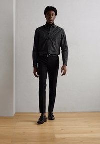 Black striped shirt with a fitted design, paired with slim black trousers and shiny black shoes, standing on a wooden floor against a light backdrop.