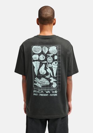 Mężczyzna w czarnej koszulce z białymi ilustracjami skamieniałości i ptaków oraz napisem „RVCA VA Past Present Future” na plecach na jednolitym tle.