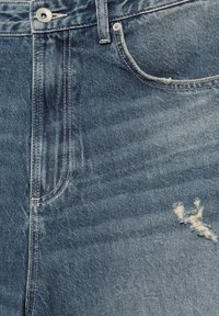 Gros plan sur un jean en denim bleu montrant un bouton en métal, une poche avant, des coutures visibles et une petite zone déchirée près de la poche.