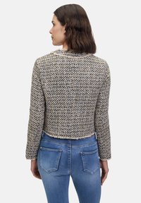 Krótka marynarka z tweedu w kolorze beżowym, czarnym i białym, z teksturowanym wzorem, postrzępionymi brzegami i długimi rękawami. Noszona na niebieskich dżinsach.